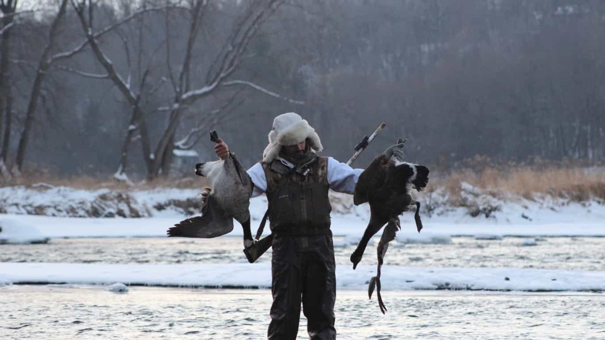 Hunter holding two harvested geese beside a snowy river after a successful goose hunt